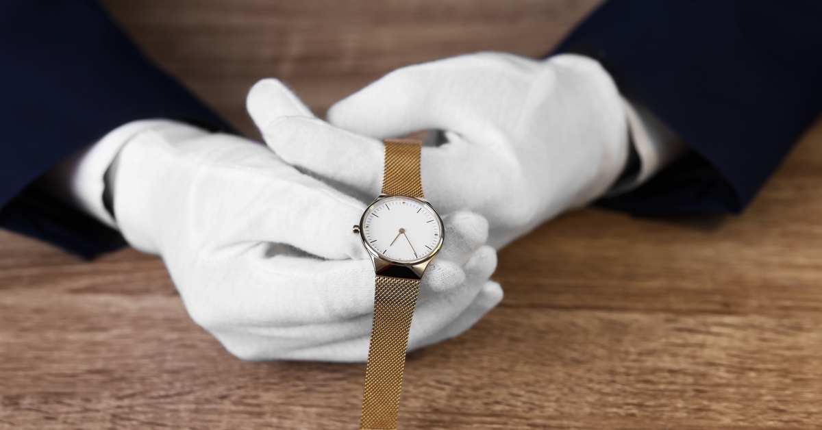 Close-up of two hands wearing white gloves holding a thin gold watch with a white face over a wooden table.
