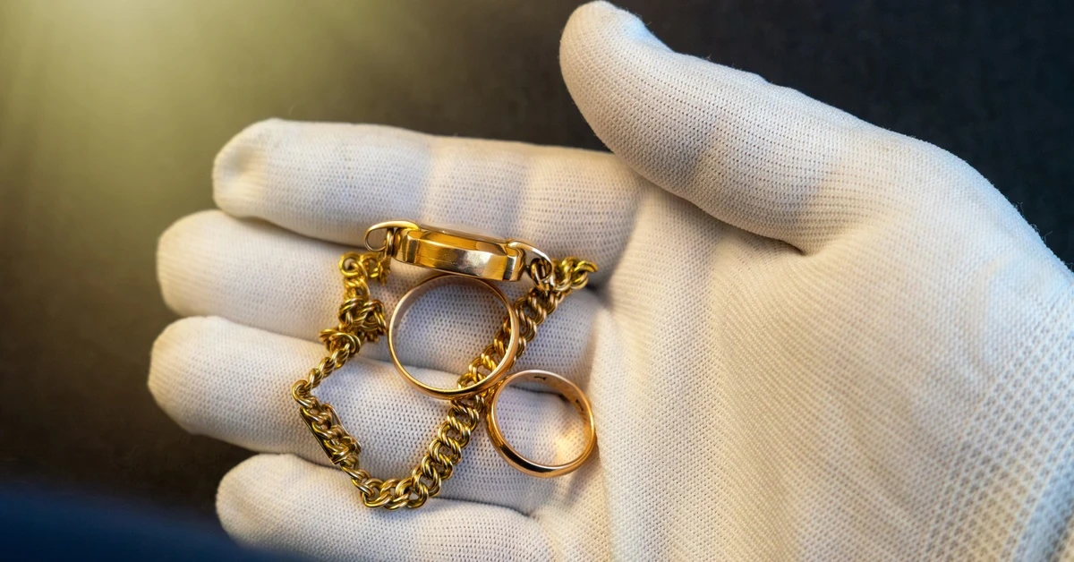 Close-up of a hand wearing a white glove holding two gold rings and a bracelet with a dim light in the background.