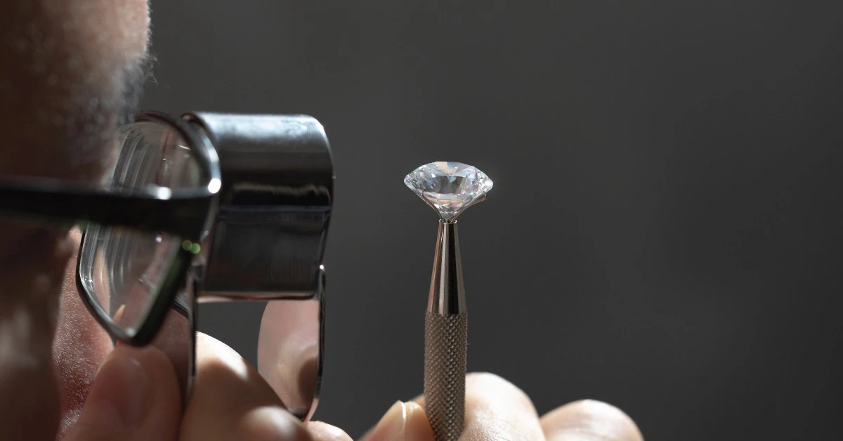 Close-up to the face of a man wearing glasses, using a metal magnifying glass to look at a small diamond on a pen.