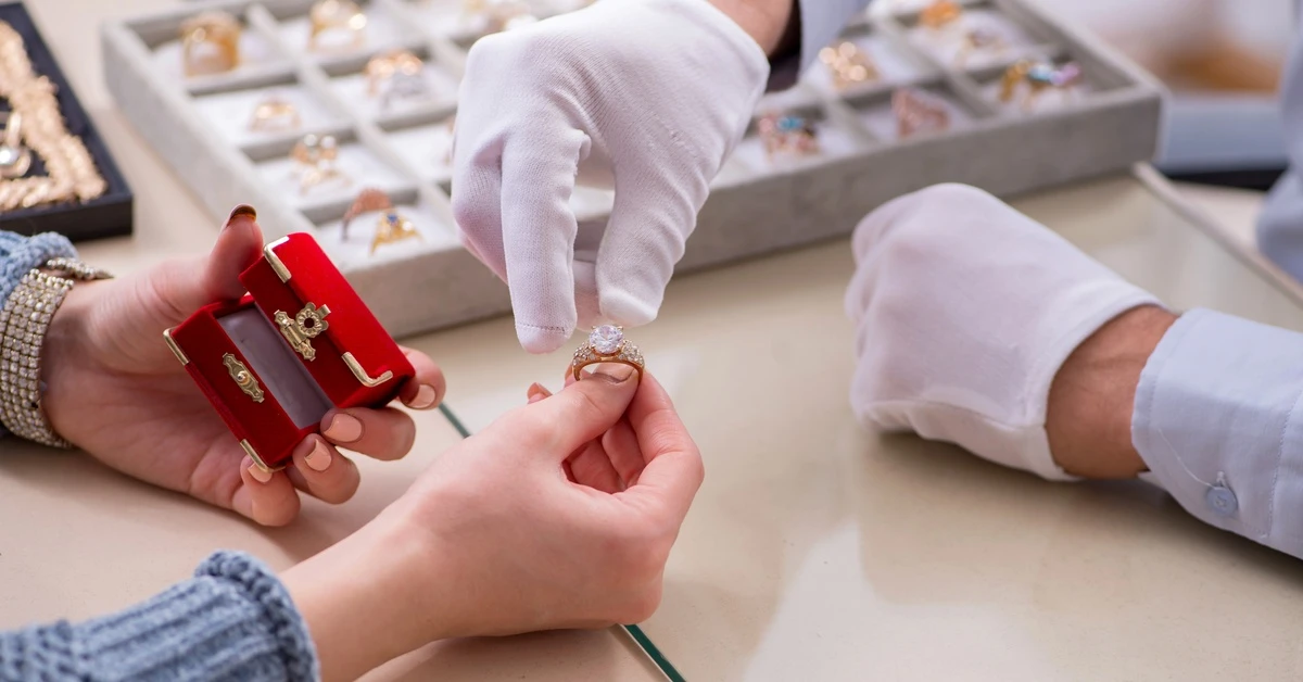 Close-up of a hand holding a large diamond ring over a counter, with a hand wearing a glove, reaching for it.